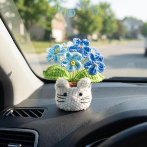 May include: A crocheted white cat-shaped planter containing several blue and light blue flowers with yellow centers and green leaves. The planter features embroidered black whiskers, two small ears, and black bead eyes on a white yarn base.