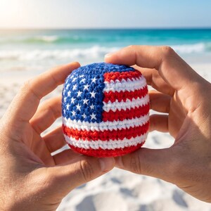 May include: A crocheted ball featuring the American flag design, with blue, white, and red stripes and stars. The ball is held in two hands against a beach backdrop with the ocean in the background.