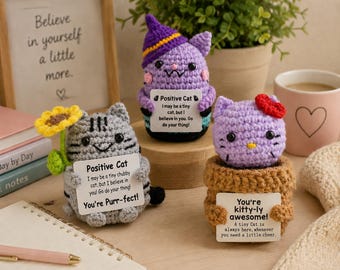 May include: Three crocheted cat figurines with positive message signs. One grey cat holds a yellow sunflower, one purple cat wears a witch hat, and one light purple cat sits in a brown pot. Each cat features black eyes and embroidered details.