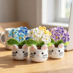 May include: Three crocheted potted plants featuring white cat-shaped containers with pink ears and black whiskers. Each pot holds a cluster of five-petaled flowers in blue, white, and yellow, or purple, with green crocheted leaves and brown soil.