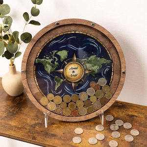 Op de afbeelding: Een ronde houten spaarpot met een transparant voorpaneel met een wereldkaartillustratie en een centraal kompasontwerp met de tekst Around the World. De binnenkant bevat diverse metalen munten, met extra munten eronder verspreid.