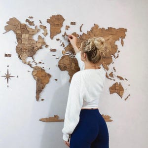 May include: A wooden world map wall decor in shades of brown is mounted on a white wall. The map is made of individual pieces, with a compass rose in the lower left corner. A person is seen interacting with the map.