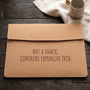 Può includere: Una custodia per laptop beige con la scritta "NOT A SNACK, CONTAINS EXPENSIVE TECH" in marrone scuro. La custodia è su una superficie di legno scuro, con una piccola tazza di ceramica e un panno di lino sullo sfondo.
