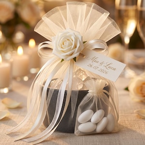 May include: A decorative wedding favor featuring a black container wrapped with white tulle and ribbon, adorned with a white rose. A small bag of white candies is attached. A tag reads "Mia & Luca 26.07.2026".