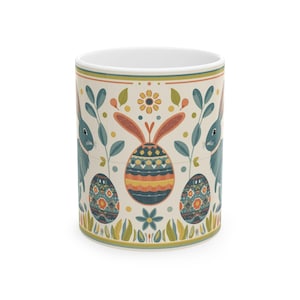 Puede incluir: Taza de cerámica blanca con un diseño de Pascua. El diseño presenta dos conejos azules, huevos de Pascua decorados y elementos florales en una paleta de colores pastel. La taza tiene forma cilíndrica y un interior blanco.