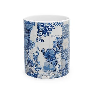 May include: White ceramic mug with a blue and white floral tile pattern. The design includes floral motifs and the word "DENMARK" in a grid-like layout. The mug has a white interior and a cylindrical shape.