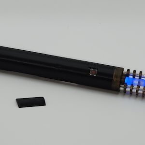 Puede incluir: Un dispositivo cilíndrico negro con un botón rectangular y una sección con luz azul. Una pequeña pieza rectangular negra se encuentra junto al dispositivo. El dispositivo tiene una apariencia metálica envejecida.