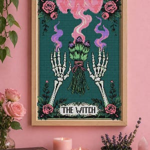 May include: Cross-stitch art in a wooden frame. The artwork features skeleton hands holding a bouquet, with pink and purple smoke rising above. The text "THE WITCH" is displayed below the bouquet, with pink roses in the corners.