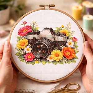 May include: Cross-stitch embroidery of a vintage camera surrounded by colorful flowers in a wooden hoop. The flowers include red, yellow, and white blooms. The camera is detailed with black and silver tones.