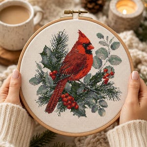 May include: A round wooden embroidery hoop containing a cross-stitch pattern of a bright red cardinal bird perched among green pine needles, holly leaves, and small red berries on a light-colored fabric background with a metal tension screw.