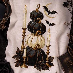 May include: A cross-stitch embroidery on white Aida fabric featuring a stack of three pumpkins in dark blue and cream colors, flanked by three tall gold candlesticks with lit candles, surrounded by dark leaves, two flying bats, and a crescent moon.