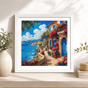 May include: A framed print of a coastal scene with a stone pathway, buildings, and vibrant flowers. The artwork features a blue sea, sky, and buildings with blue doors and shutters. The print is in a white frame.