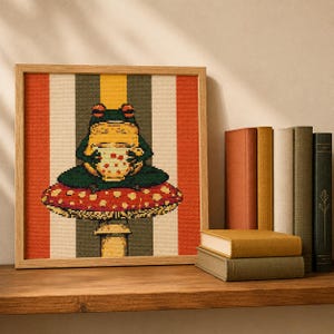May include: A square framed cross-stitch embroidery featuring a green frog sitting on a red and white spotted mushroom while holding a white mug with red dots. The background consists of vertical stripes in orange, yellow, and dark gray.