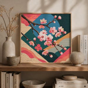 May include: A framed cross-stitch artwork featuring a branch of pink cherry blossoms against a geometric background of teal, blue, and orange. The artwork is displayed on a wooden shelf with decorative items.