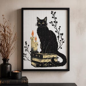 Op de afbeelding: Kruissteekkunst van een zwarte kat die op een stapel boeken zit, met twee brandende kaarsen en takjes. De kat heeft gele ogen en is omlijst in een zwarte rand. Het kunstwerk wordt op een witte achtergrond getoond.