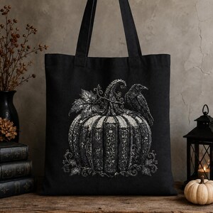 May include: A black tote bag featuring a detailed graphic print of a pumpkin constructed from stacked vintage books. A raven is perched on top of the pumpkin, which is surrounded by decorative leaves and vines. The design is rendered in shades of grey.