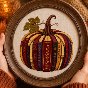 May include: A circular cross-stitch embroidery featuring a pumpkin shape constructed from vertical book spines in shades of deep red, purple, and gold. The design includes a green leaf and a brown stem, all encased in a dark brown wooden frame.