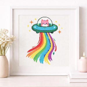 May include: Cross-stitch art featuring a pink cat in a spaceship, emitting a rainbow. The spaceship is teal with a clear dome. Yellow stars surround the craft. The artwork is framed in a white square frame.