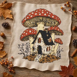 May include: A square piece of beige fabric featuring a cross-stitch embroidery of a house nestled beneath large red mushrooms with white spots. The house has a black roof, white walls, and a dark arched doorway, surrounded by small bare branches.