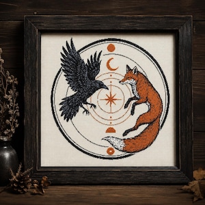 May include: A framed cross-stitch embroidery featuring a black raven and an orange fox facing each other within a circular geometric design. The composition includes celestial symbols like a sun, moon, and star, all rendered in black and orange thread.