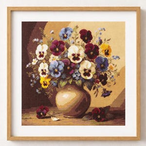 May include: A framed cross-stitch artwork featuring a still life of colorful pansies in a vase. The pansies are in shades of white, purple, blue, and yellow. The vase is a light brown color, and the background is a gradient of brown and beige.
