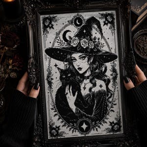 May include: A black and white cross-stitch embroidery featuring a person wearing a wide-brimmed witch hat adorned with roses, holding a black cat. The design is framed in an ornate black rectangular frame with intricate decorative carvings.