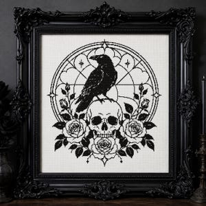 May include: A black and white cross-stitch embroidery featuring a raven perched atop a human skull, surrounded by roses and set within a circular geometric frame. The artwork is displayed inside a decorative, ornate black rectangular frame.