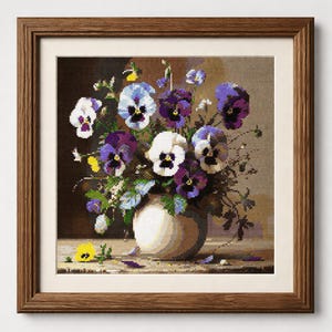 May include: A cross-stitch embroidery piece framed in wood, depicting a bouquet of pansies in shades of purple, white, and yellow arranged in a rounded white vase. The flowers are set against a dark background, with a single yellow pansy resting on the surface.