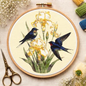 May include: Cross-stitch embroidery of two blue birds perched near yellow irises. The embroidery is set in a wooden hoop. The birds have dark blue feathers with red accents. The irises have yellow petals and green leaves. The background is a light cream color.