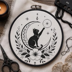 May include: A circular embroidery hoop containing a black cross-stitch design on white fabric. The pattern features a black cat sitting on a crescent moon, reaching for a star, surrounded by floral motifs, small stars, and celestial symbols.