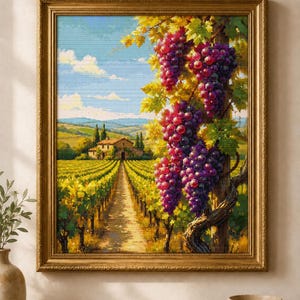 Pode incluir: Um bordado em ponto cruz emoldurado retratando uma paisagem de vinhedo com um caminho de terra central que leva a uma casa de pedra sob um céu azul. Em primeiro plano, grandes cachos de uvas roxas escuras pendem de um poste de madeira.
