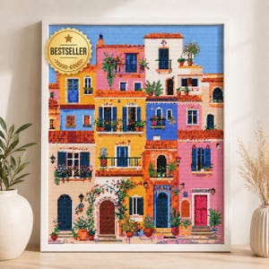 Op de afbeelding: Een ingelijst kruissteek- of diamond painting-kunstwerk met een kleurrijke mediterrane straatscène met gebouwen van meerdere verdiepingen in roze, geel en blauw, voorzien van balkons, potplanten en een gouden BESTSELLER-zegel.