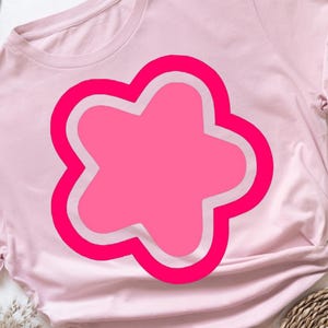 May include: A light pink t-shirt featuring a large, bright pink flower design with a white outline. The flower has a rounded, four-petal shape. The shirt is made of soft material and has short sleeves.