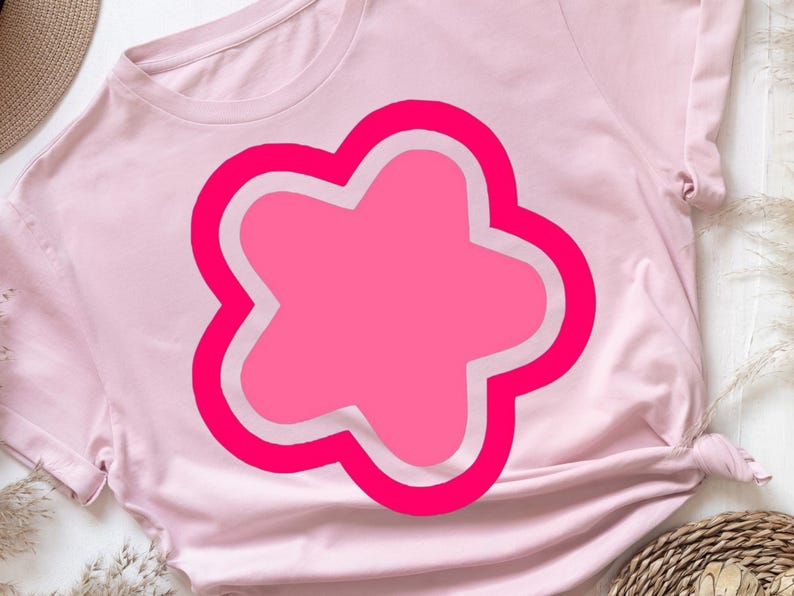 May include: A light pink t-shirt featuring a large, bright pink flower design with a white outline. The flower has a rounded, four-petal shape. The shirt is made of soft material and has short sleeves.
