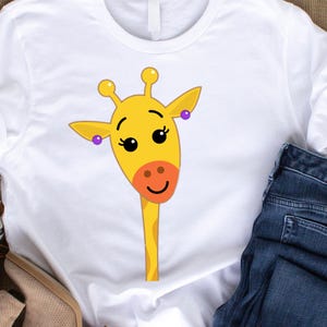 May include: White t-shirt featuring a cartoon giraffe face. The giraffe has a yellow head, orange nose, and purple earrings. The shirt is paired with blue jeans and tan shoes.