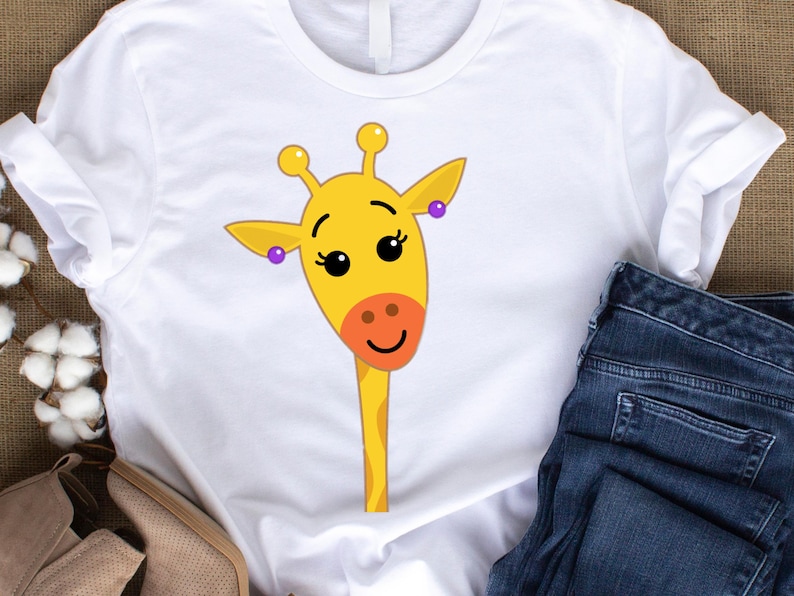 May include: White t-shirt featuring a cartoon giraffe face. The giraffe has a yellow head, orange nose, and purple earrings. The shirt is paired with blue jeans and tan shoes.