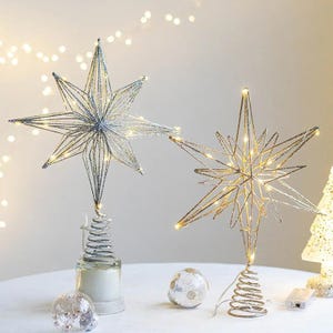 Könnte beinhalten: Zwei sternförmige Weihnachtsbaumspitzen mit glitzernden silbernen und goldenen Drahtrahmen. Eine hat eine silberne Oberfläche, die andere gold, beide mit warmweißen Lichtern. Jeder Stern sitzt auf einem dekorativen Sockel, perfekt für die Weihnachtsdekoration.