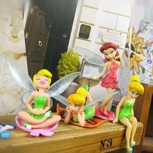 K&ouml;nnte beinhalten: Eine Sammlung von Feenfiguren, jede mit einzigartigen Posen und Outfits. Die Feen haben farbenfrohe Kleider, Fl&uuml;gel und Frisuren. Die Figuren sind auf einer Holzoberfl&auml;che ausgestellt und schaffen eine skurrile Szene.