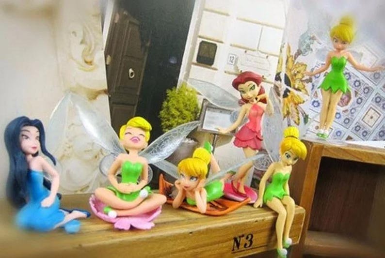 K&ouml;nnte beinhalten: Eine Sammlung von Feenfiguren, jede mit einzigartigen Posen und Outfits. Die Feen haben farbenfrohe Kleider, Fl&uuml;gel und Frisuren. Die Figuren sind auf einer Holzoberfl&auml;che ausgestellt und schaffen eine skurrile Szene.