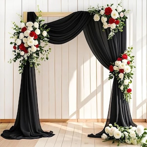 May include: A rectangular wooden wedding arch draped with flowing black fabric. The structure is decorated with three large floral arrangements consisting of white and red roses, along with green foliage, positioned on the top right and sides.