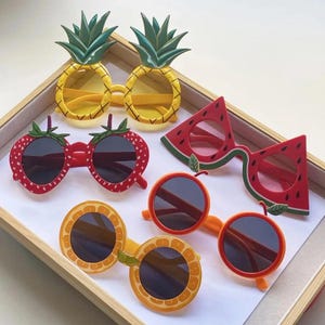 May include: A collection of five pairs of novelty sunglasses shaped like fruit. The set includes pineapple-shaped glasses with yellow lenses, strawberry-shaped glasses, watermelon slice-shaped glasses, and two pairs of orange slice-shaped glasses.
