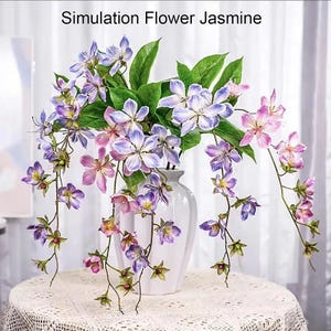 May include: A white vase holding artificial jasmine flowers. The flowers are a blend of purple, pink, and white, with green leaves. The text "Simulation Flower Jasmine" is at the top. The vase is on a white lace tablecloth.