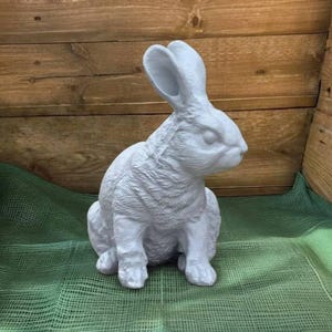 May include: A light gray rabbit statue in a seated position with its ears perked up. The bunny has detailed fur texture and is set against a wooden background and green fabric.