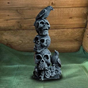May include: A stacked skull sculpture with two ravens and roses. The gray sculpture features three skulls, with a raven perched on top and another on the side. Roses are at the base, creating a gothic and spooky aesthetic.