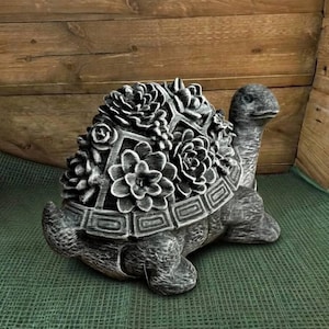 May include: A gray decorative turtle statue with a shell adorned with sculpted flowers. The statue is made of a stone-like material and has a detailed, textured surface. The turtle is positioned on a green surface.