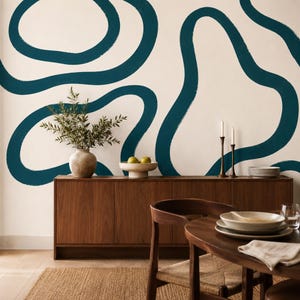 Könnte beinhalten: Eine Wand mit cremefarbenem Hintergrund und großen, dunkel-petrolfarbenen abstrakten Pinselstrichformen. Ein hölzernes Sideboard trägt eine Keramikvase mit grünen Zweigen, eine Schale mit zwei Birnen, zwei hohe Kerzenhalter und weiße Teller.