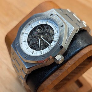 May include: A silver wristwatch with a skeleton dial, revealing the internal mechanisms. The watch has a white face with black detailing and the brand name "SEIKO". It features a stainless steel bracelet and is presented on a wooden stand.