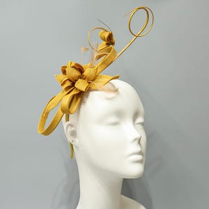 Pode incluir: Acessório de cabeça amarelo mostarda com um detalhe floral e redemoinhos decorativos. O acessório de cabeça é feito de material tecido e apresenta penas delicadas. O acessório de cabeça é usado em uma cabeça de manequim.