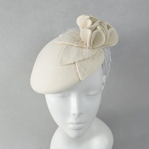May include: A cream-colored felt fascinator hat featuring a structured, layered floral accent on the side. The headpiece is adorned with a delicate, fine-mesh netting veil that drapes over the top, providing a textured and layered appearance.