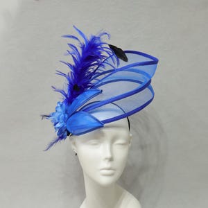 Könnte beinhalten: Ein leuchtend königsblauer Fascinator mit einem strukturierten, mehrschichtigen Netzdesign. Das Kopfstück ist mit einer Gruppe passender blauer Stoffblumen und langen, feinen Federn verziert, die nach oben ragen und eine dynamische Silhouette bilden.